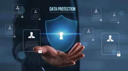 A person in a business suit holds out their hand, above which a glowing digital shield with a keyhole icon appears, symbolizing security. Surrounding the shield are floating profile icons with small padlocks, representing protected personal data. The words “Data Protection” are displayed above the shield, emphasizing cybersecurity and privacy